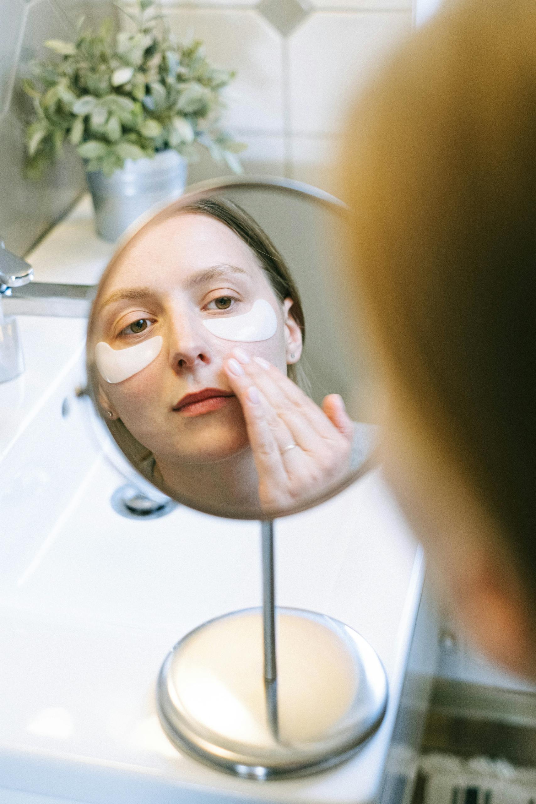 Woman applying eye patches as part of her skincare routine, reflected in bathroom mirror.